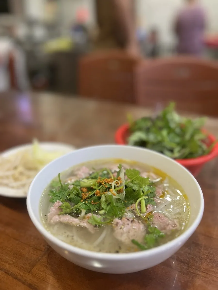 A bowl of Hoi An beef pho served with bean sprouts and a basket of fresh herbs.