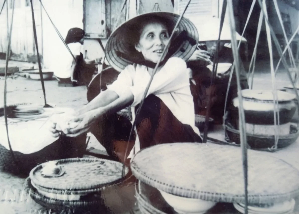 A woman with a bamboo carrying pole selling mi quang from a street corner in Hoi An, surrounded by small plastic stools and customers