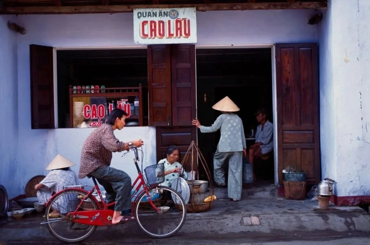 Black and white photo from 1990 showing a woman selling bun on the doorstep of a house in Hoi An, with a cao lau shop visible behind her