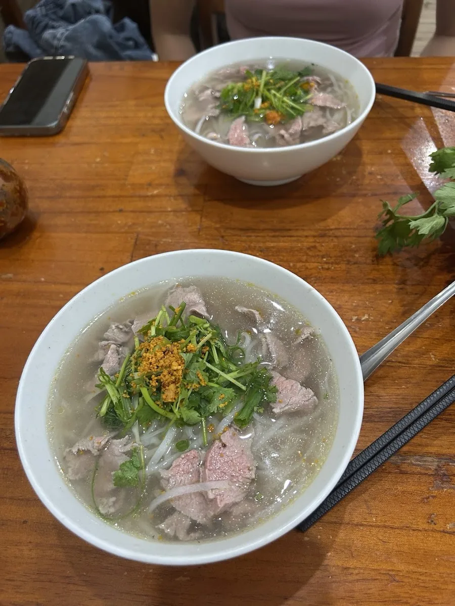 Two bowls of Hoi An beef pho on a wooden table with rare beef slices, cilantro and fried shallots