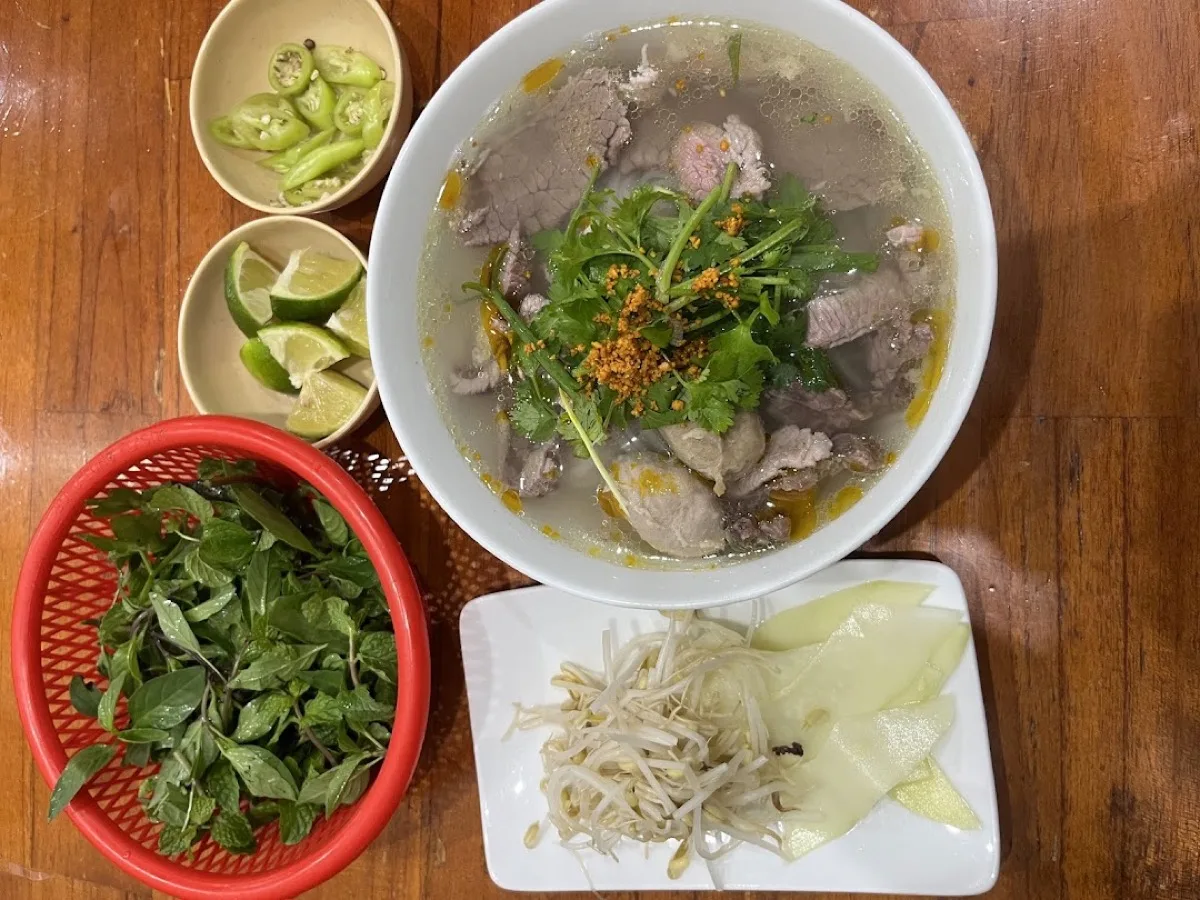 A bowl of Hoi An pho with the full table spread — clear broth, beef slices, fresh herb basket, bean sprouts, lime and chili ready for the diner to season