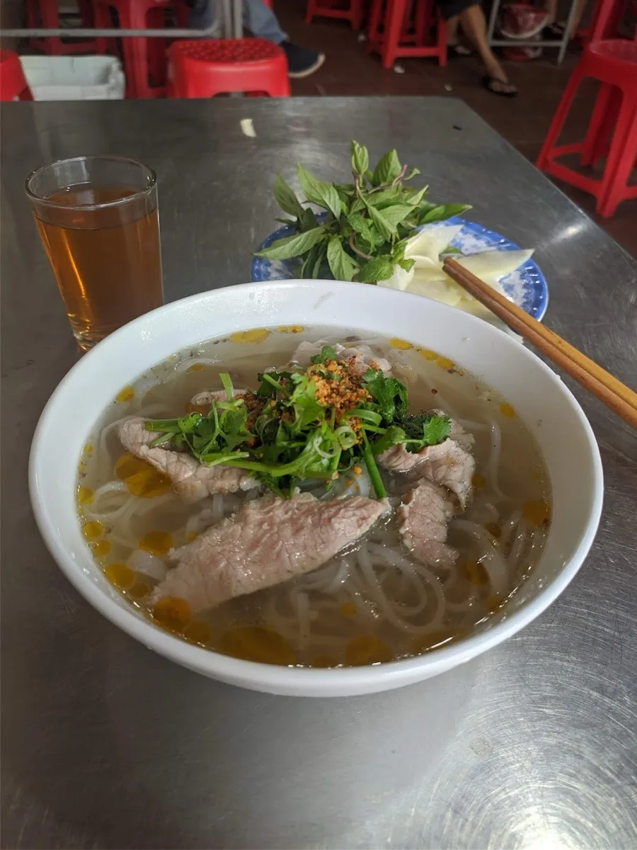 A bowl of beef pho with iced tea and a plate of fresh herbs at a Hoi An sidewalk table
