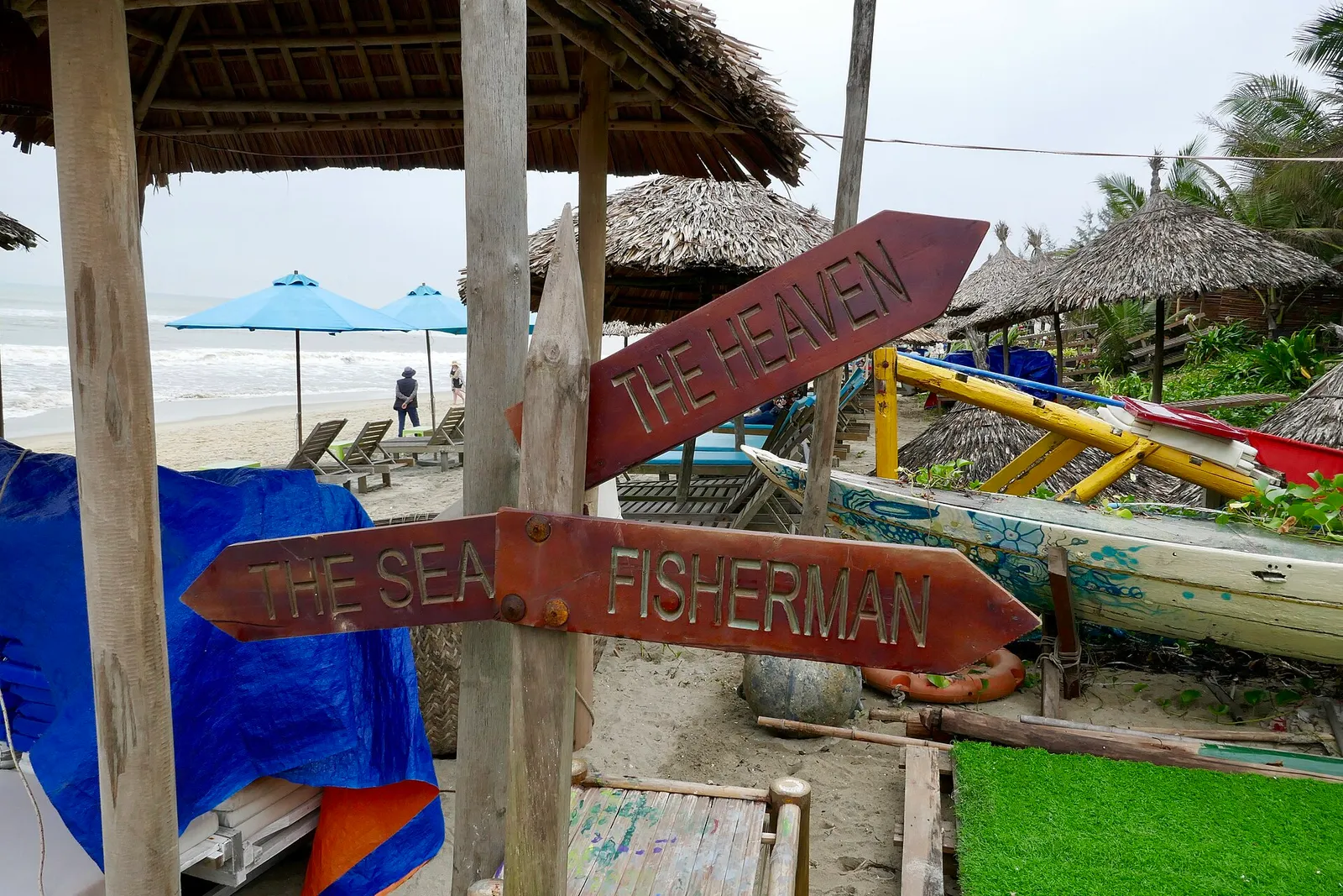 Wooden signposts and thatched umbrellas at a beach bar on An Bang beach, with fishing boats behind.