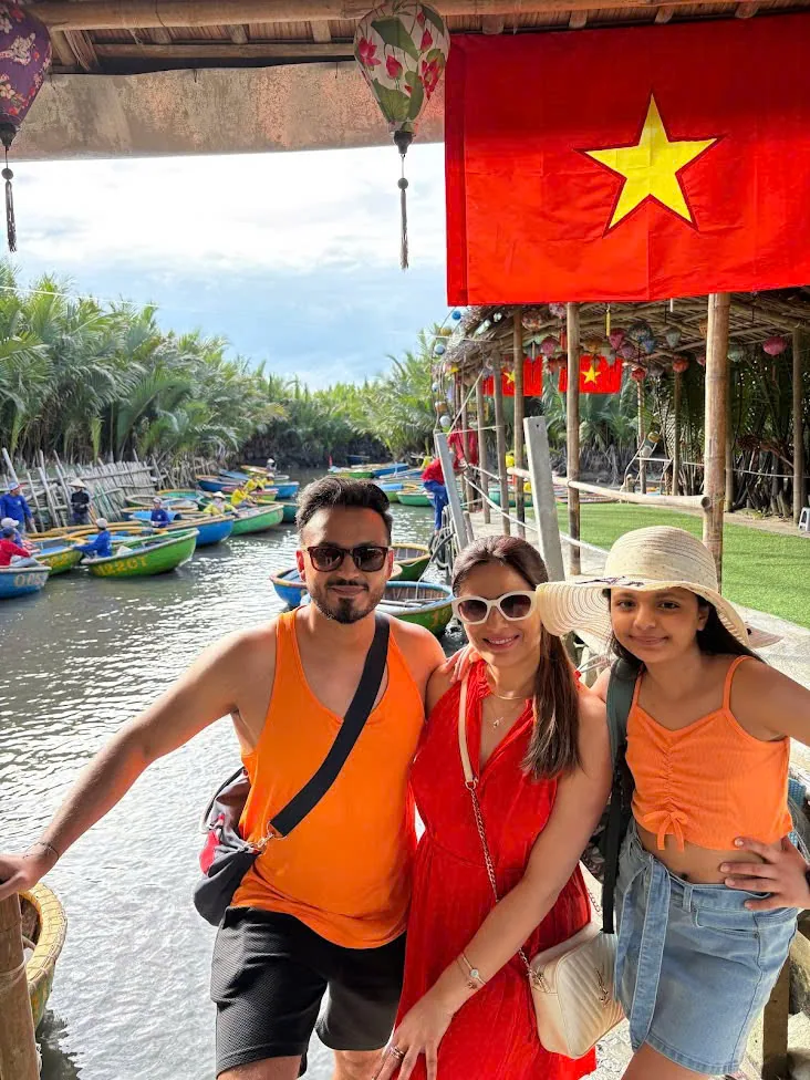 A family of three at the basket boat landing in the Cam Thanh coconut forest, under a Vietnamese flag.