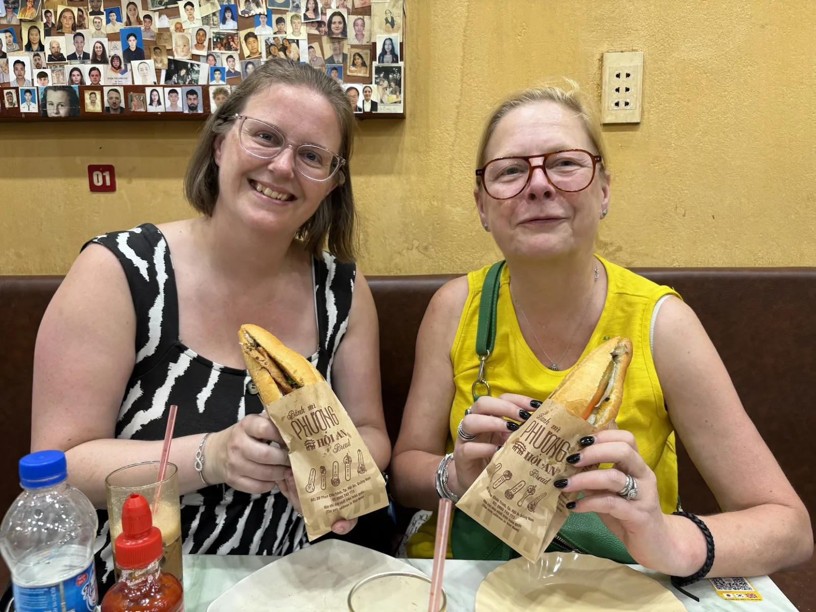 Two guests sitting at Banh Mi Phuong in Hoi An holding banh mi in paper sleeves, a wall of photographs behind them.