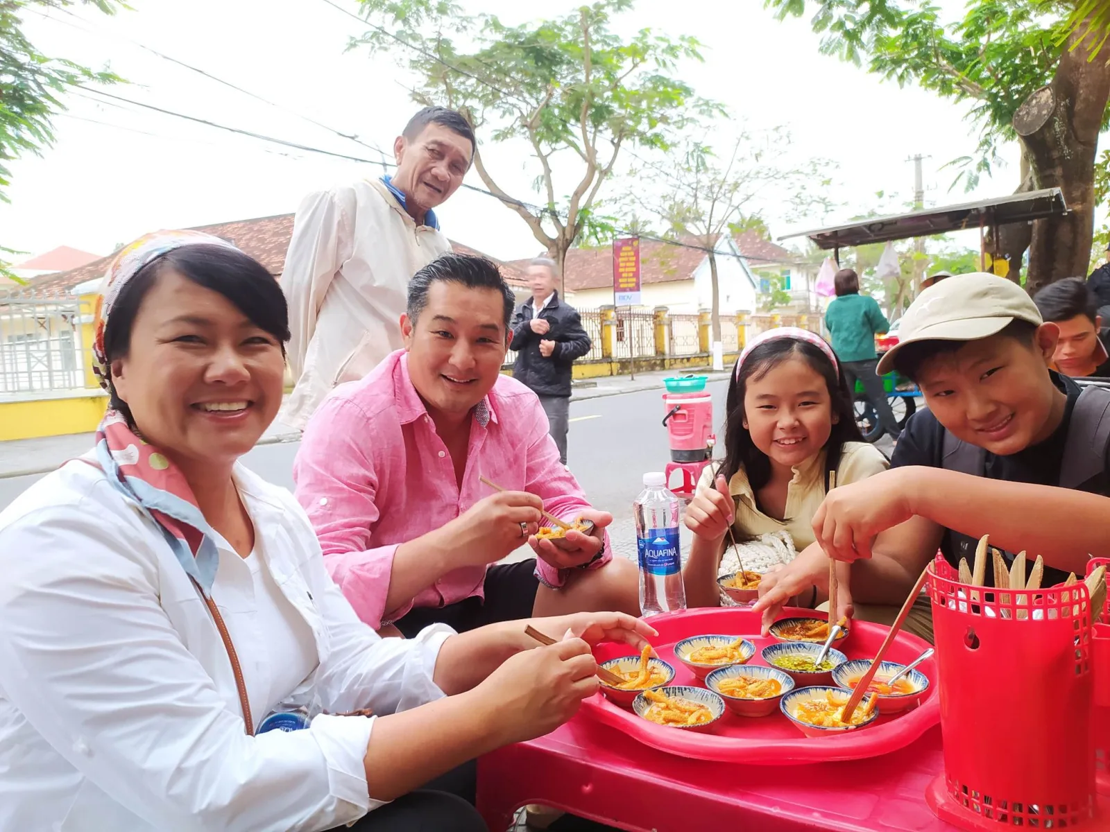A family eating small bowls of a street snack at a red plastic table beside a food cart on a Hoi An street.
