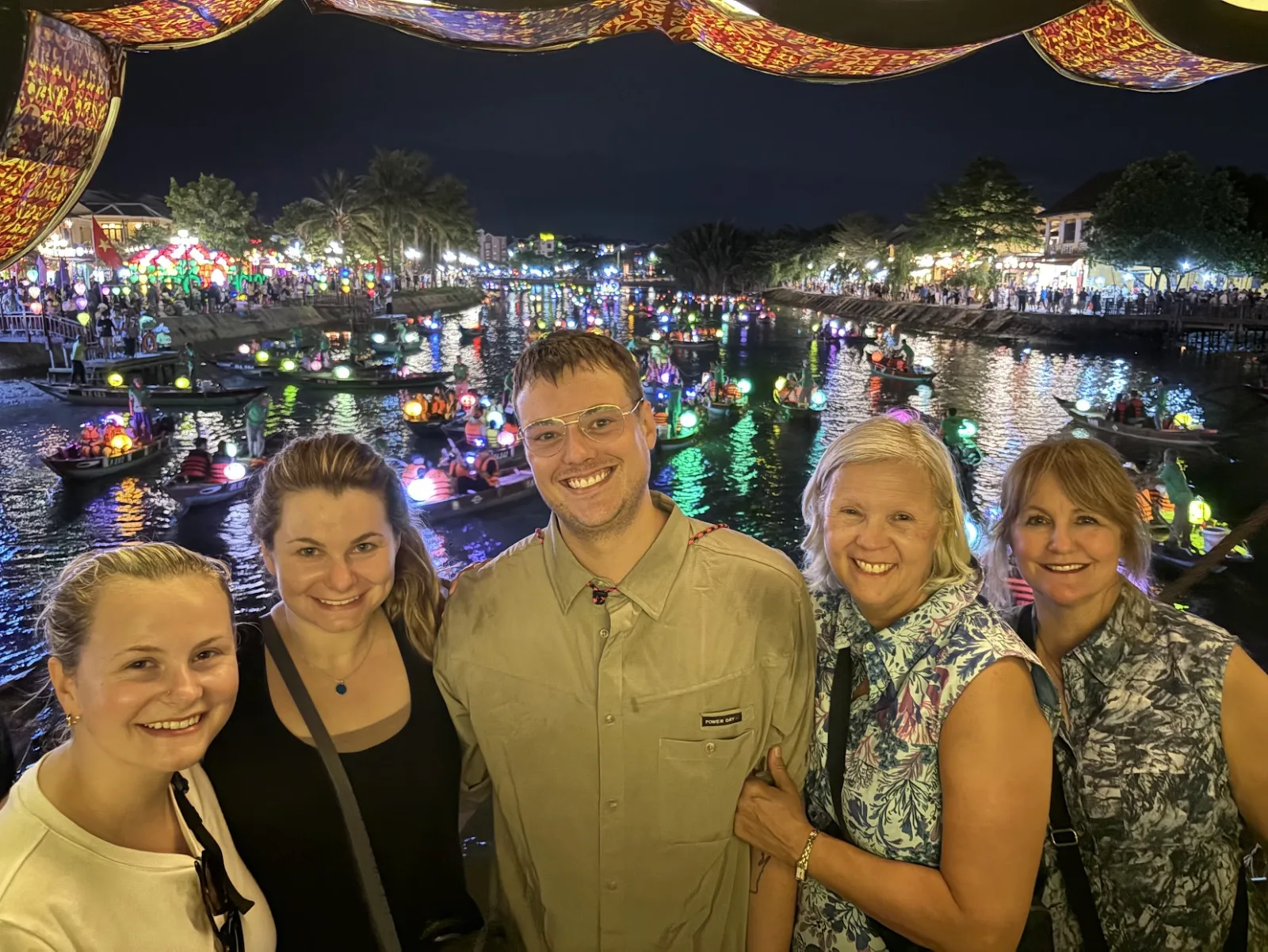 Private tour group by the Hoi An riverside at night, with lantern-lit boats drifting on the Thu Bon behind them.