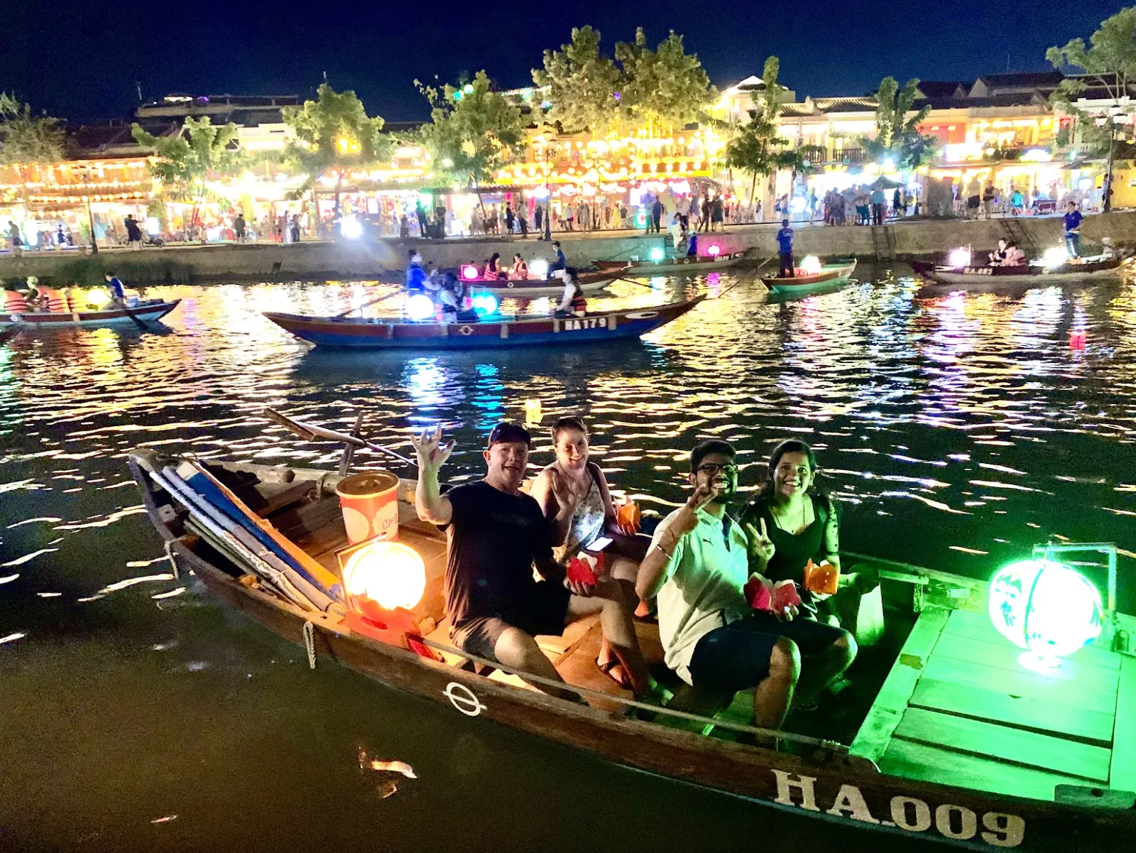 Guests on a wooden boat holding glowing lanterns on the Hoai River at night.
