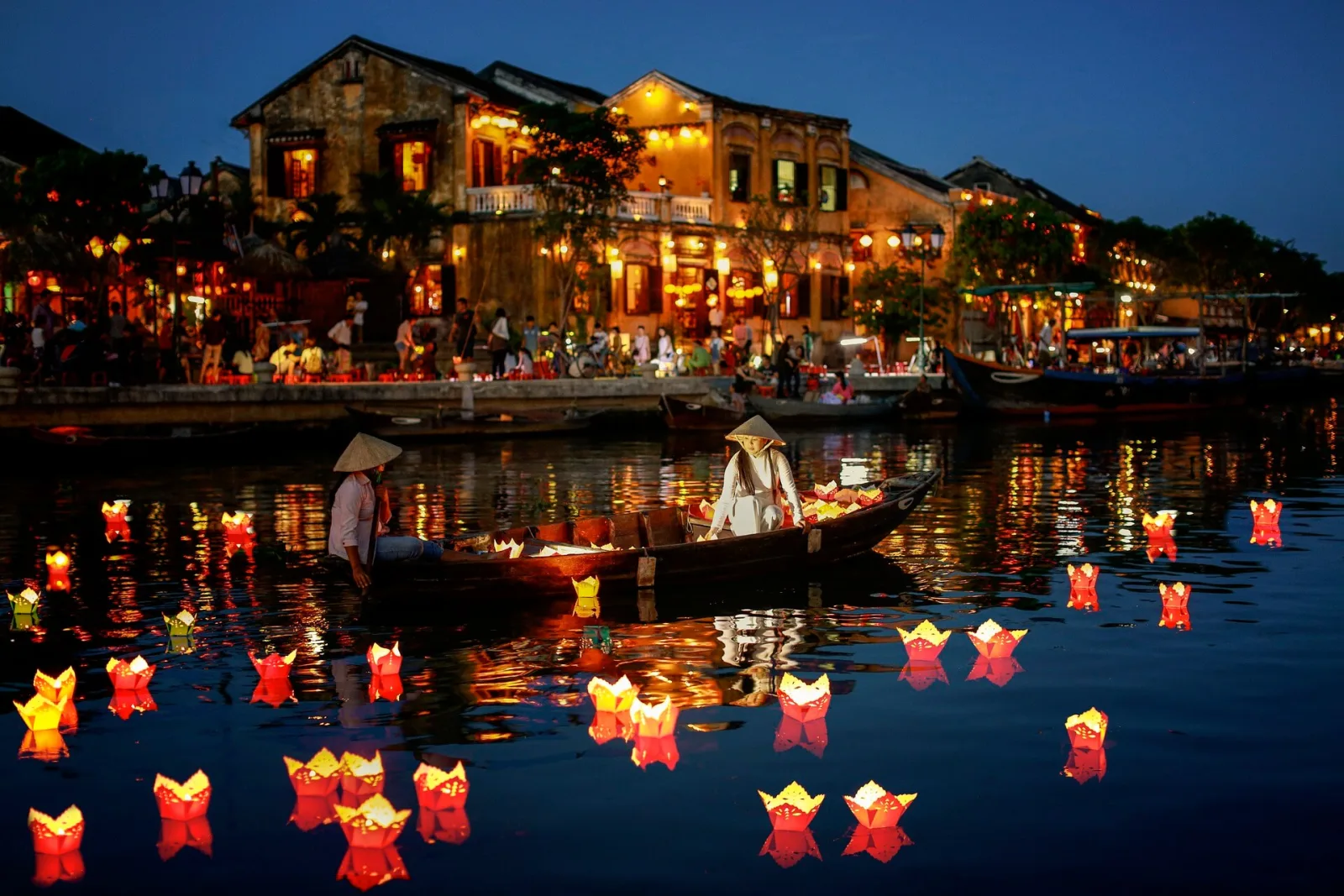 A conical-hat rower steering a lantern-lit boat past the floating lights of the Hoai River.