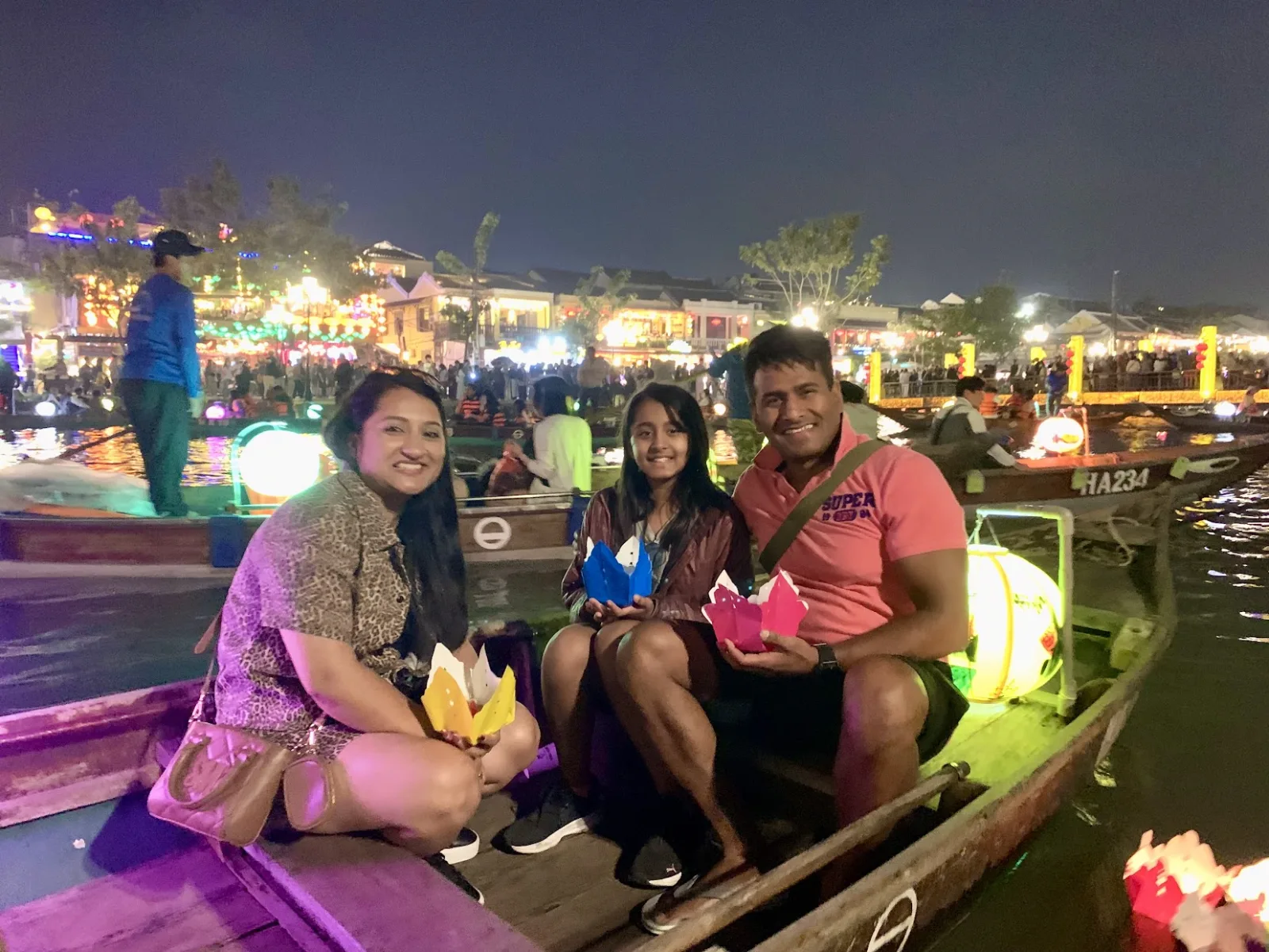 Three guests holding paper lotus lanterns on a boat with the Hoi An night market behind them.