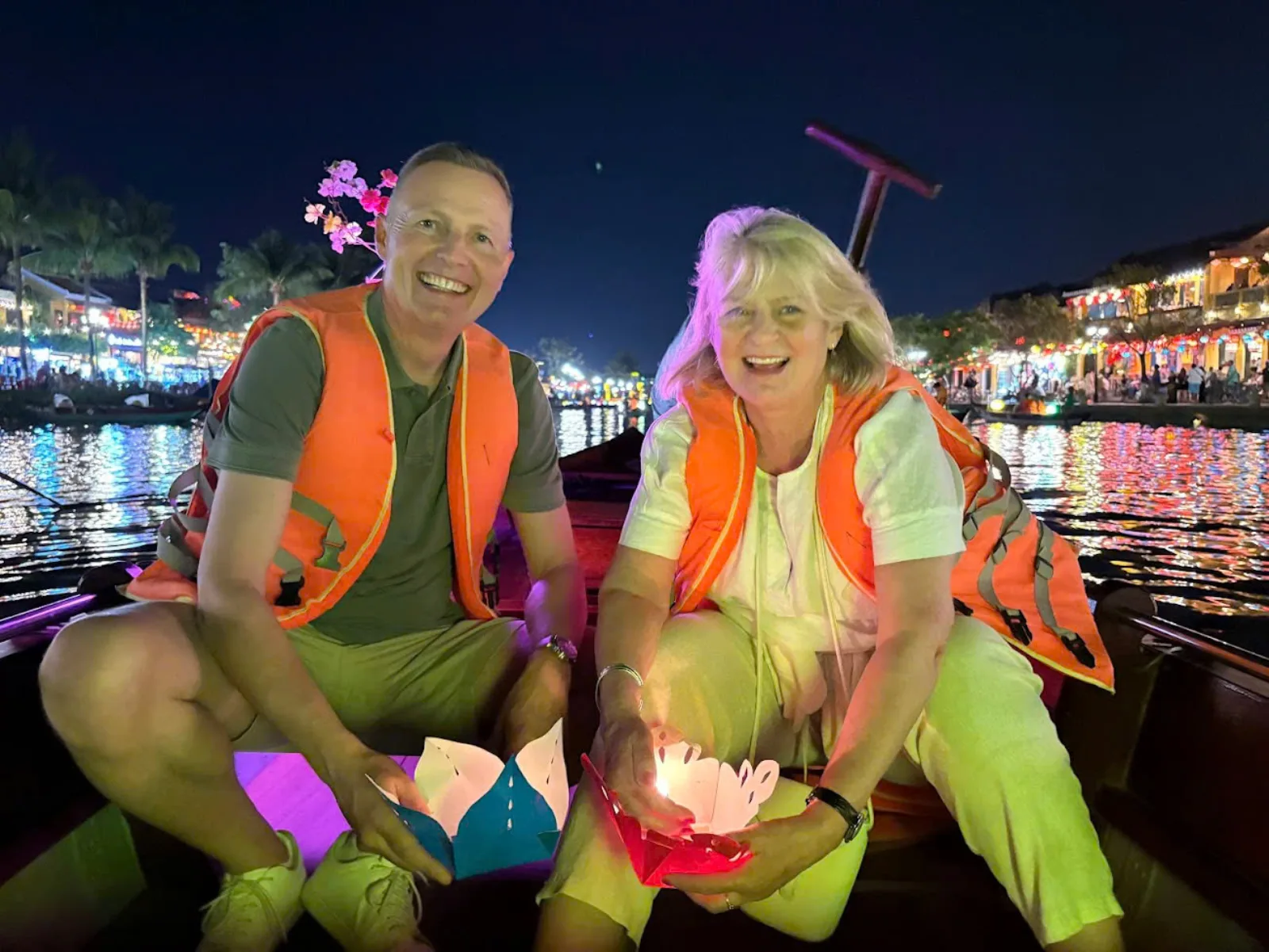 A smiling couple in life jackets holding lotus flower lanterns on a night boat in Hoi An.
