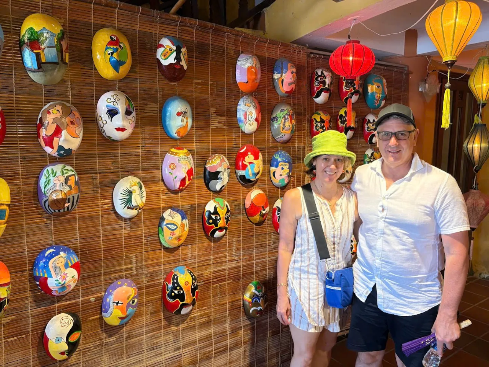 A couple photographed beside a bamboo wall of hand-painted masks and glowing lanterns.